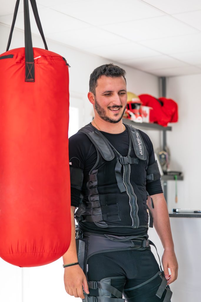 Client souriant en gilet EMS devant le sac de frappe rouge